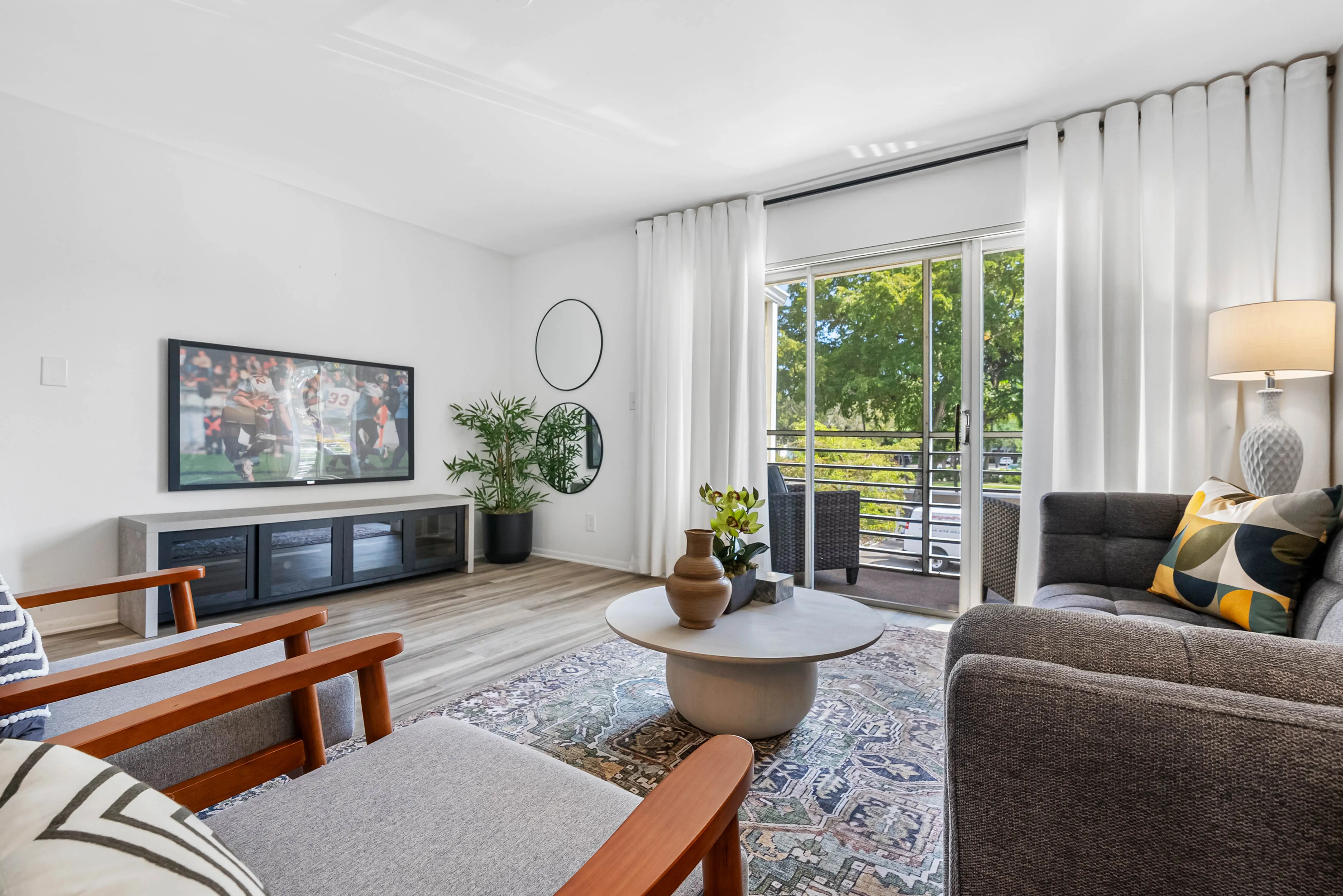 A modern living room with a combination of a dark grey sofa and wooden armchairs, a round coffee table, and a large window with curtains. A flat-screen TV is mounted on the wall, and there are plants and decorative items present.