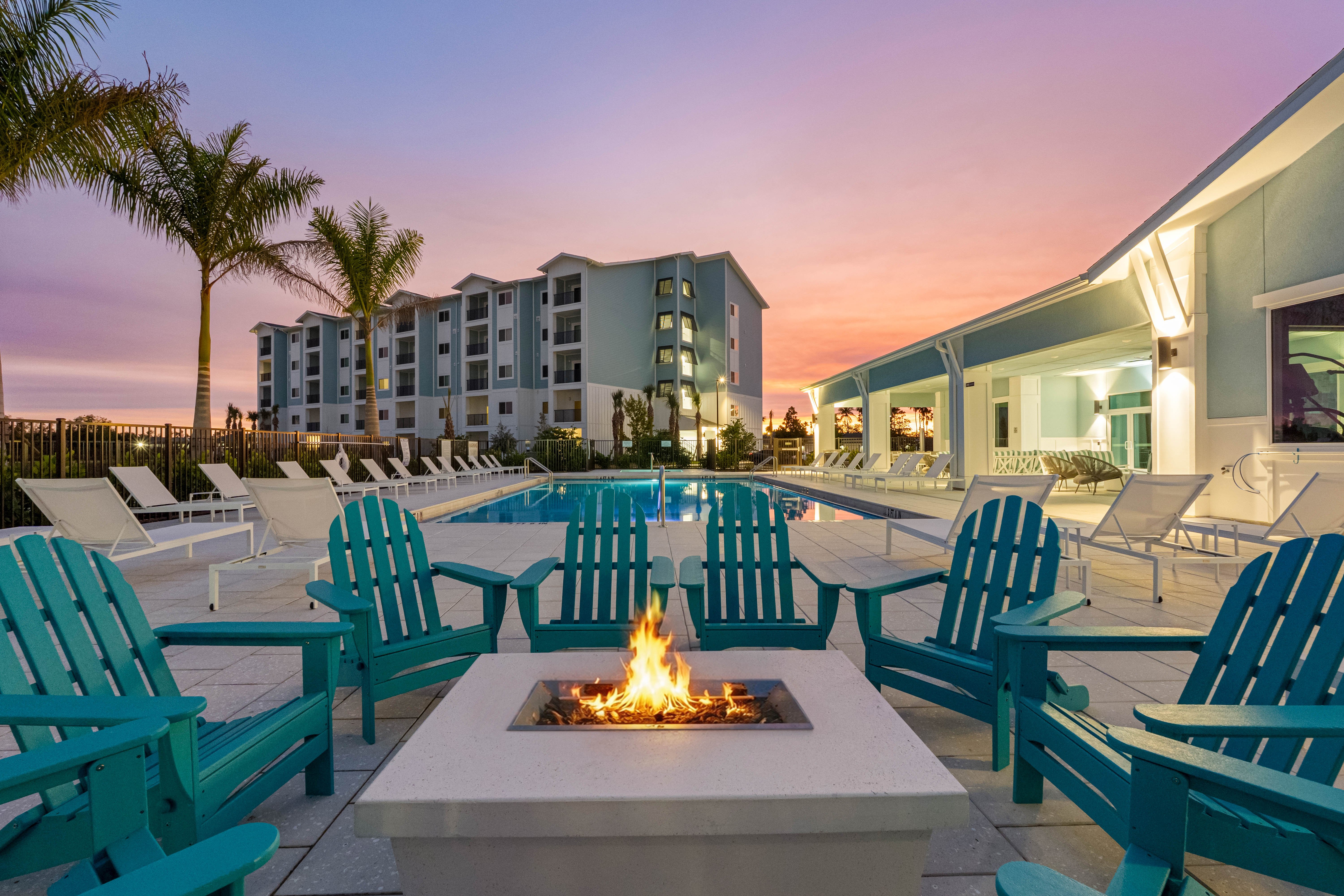 A beautiful outdoor area featuring a fire pit surrounded by turquoise chairs, with a swimming pool and a building in the background during sunset.
