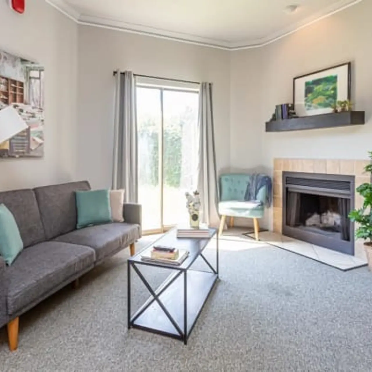 A modern living room featuring a gray sofa with teal and cream pillows, a glass coffee table, and a corner plant. The room has a fireplace, artwork on the wall, and large windows with sheer curtains allowing light in.