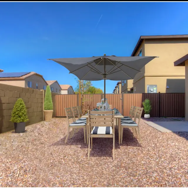 A backyard dining area features a wooden table with chairs under a large umbrella, surrounded by gravel and small plants.
