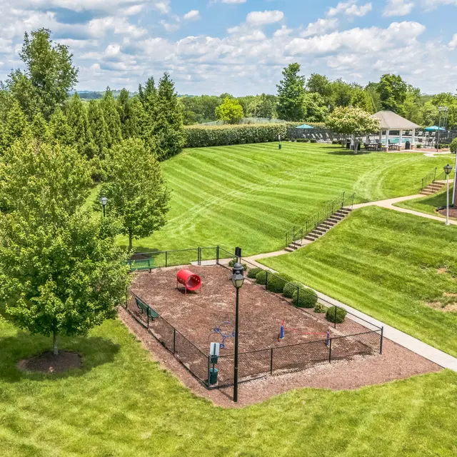 Aerial View of Park with Dog Playground An aerial view of a well-maintained park featuring a grassy area, a dog park with a red structure, trees, and a concrete path that leads to a multi-story building in the background.