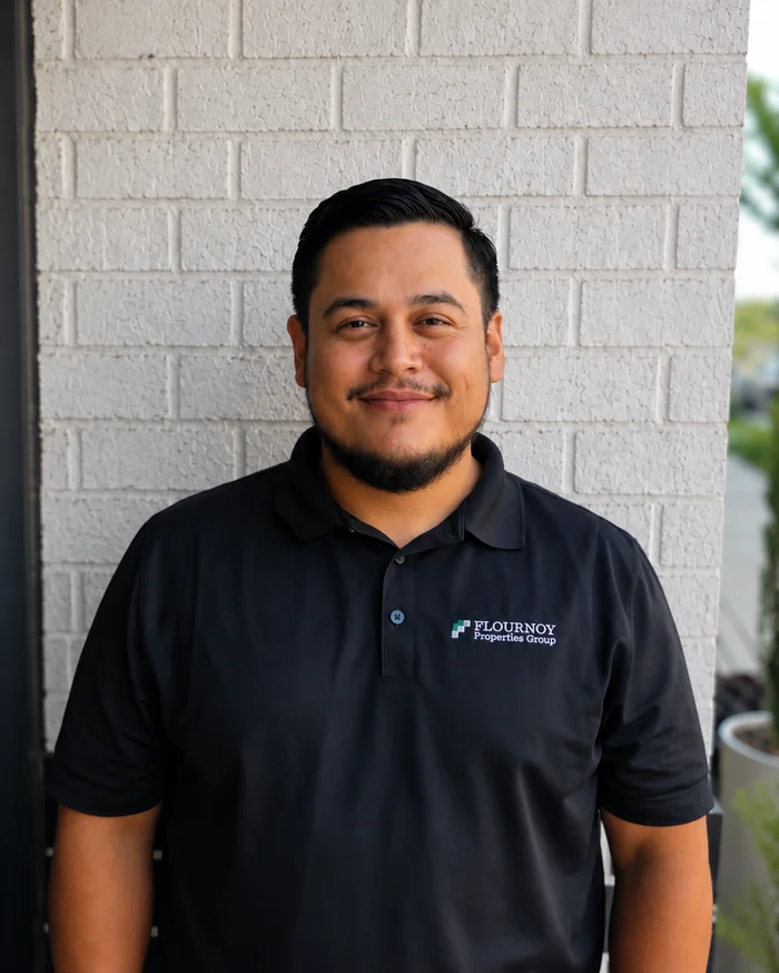 Professional Portrait A man wearing a black polo shirt with a logo stands against a brick wall. The background is blurred, showing outdoor elements such as plants.