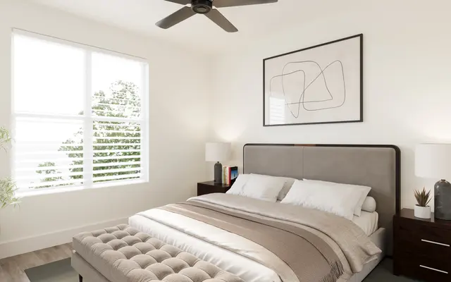A modern bedroom featuring a large bed with a grey upholstered headboard, beige bedding, and two bedside tables with lamps. There's a window with white shutters letting in natural light, and a decorative plant in the corner.