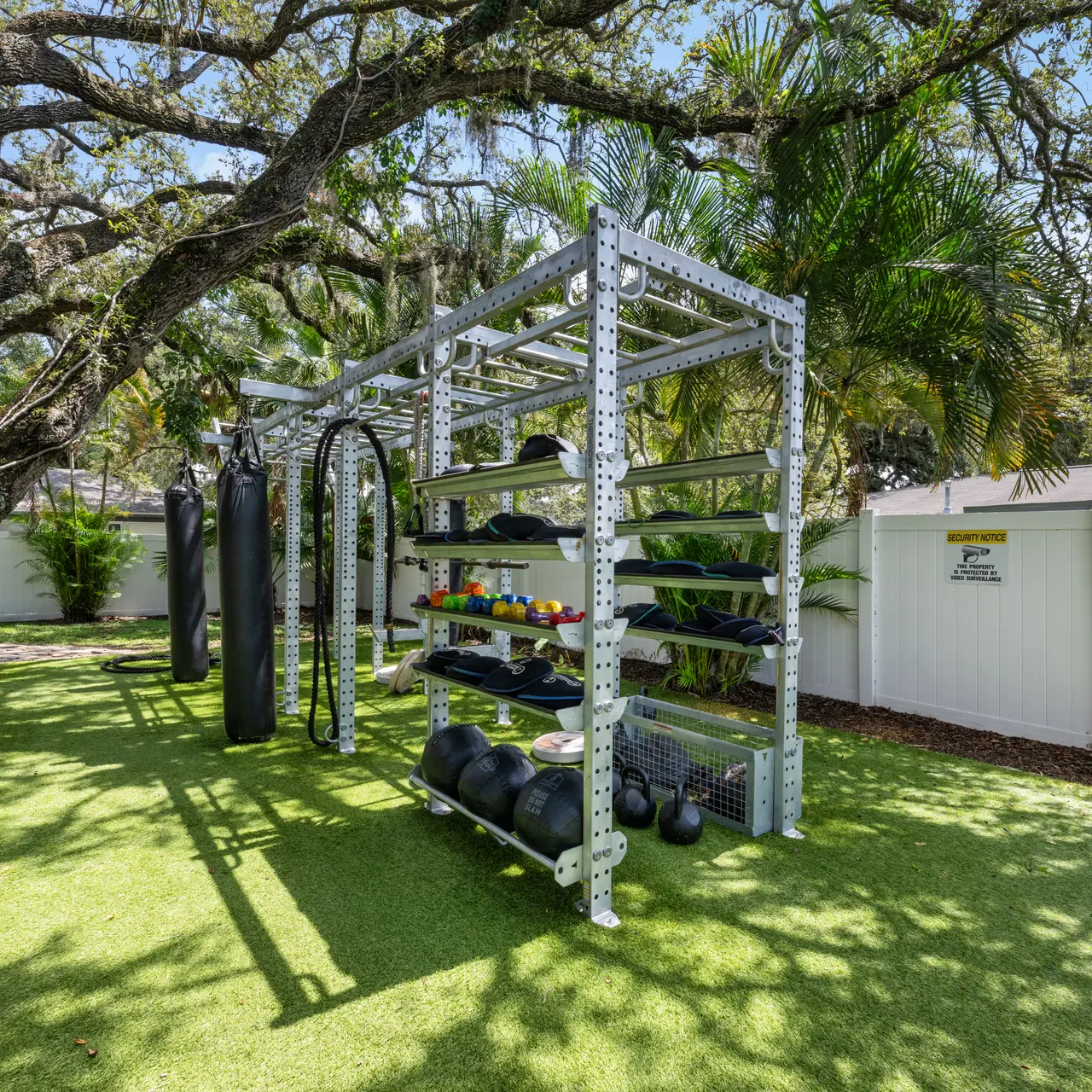 An outdoor gym setup featuring a gray metal structure with exercise equipment, including free weights and punching bags, surrounded by green grass and trees.