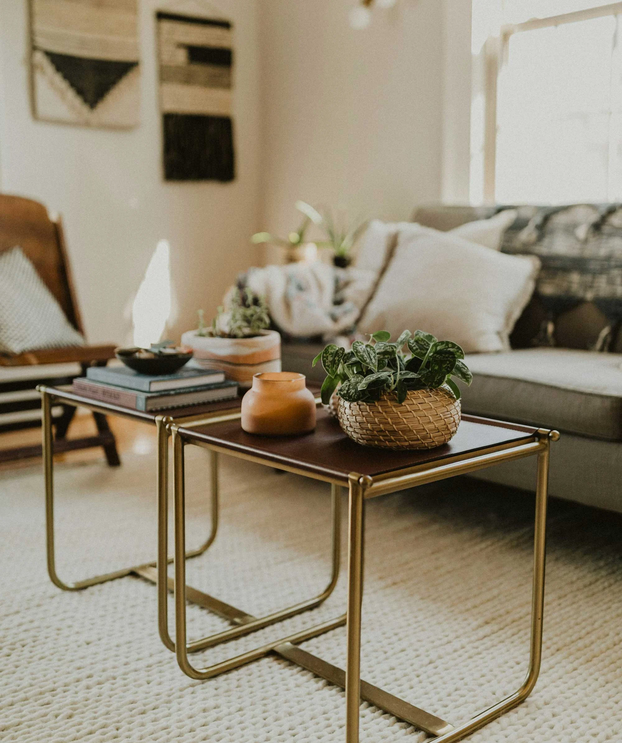 A cozy living room setup featuring two stylish side tables. One table has a stack of books and a bowl, while the other holds a plant in a woven basket and a decorative candle. A comfortable sofa and textured decor are visible in the background.