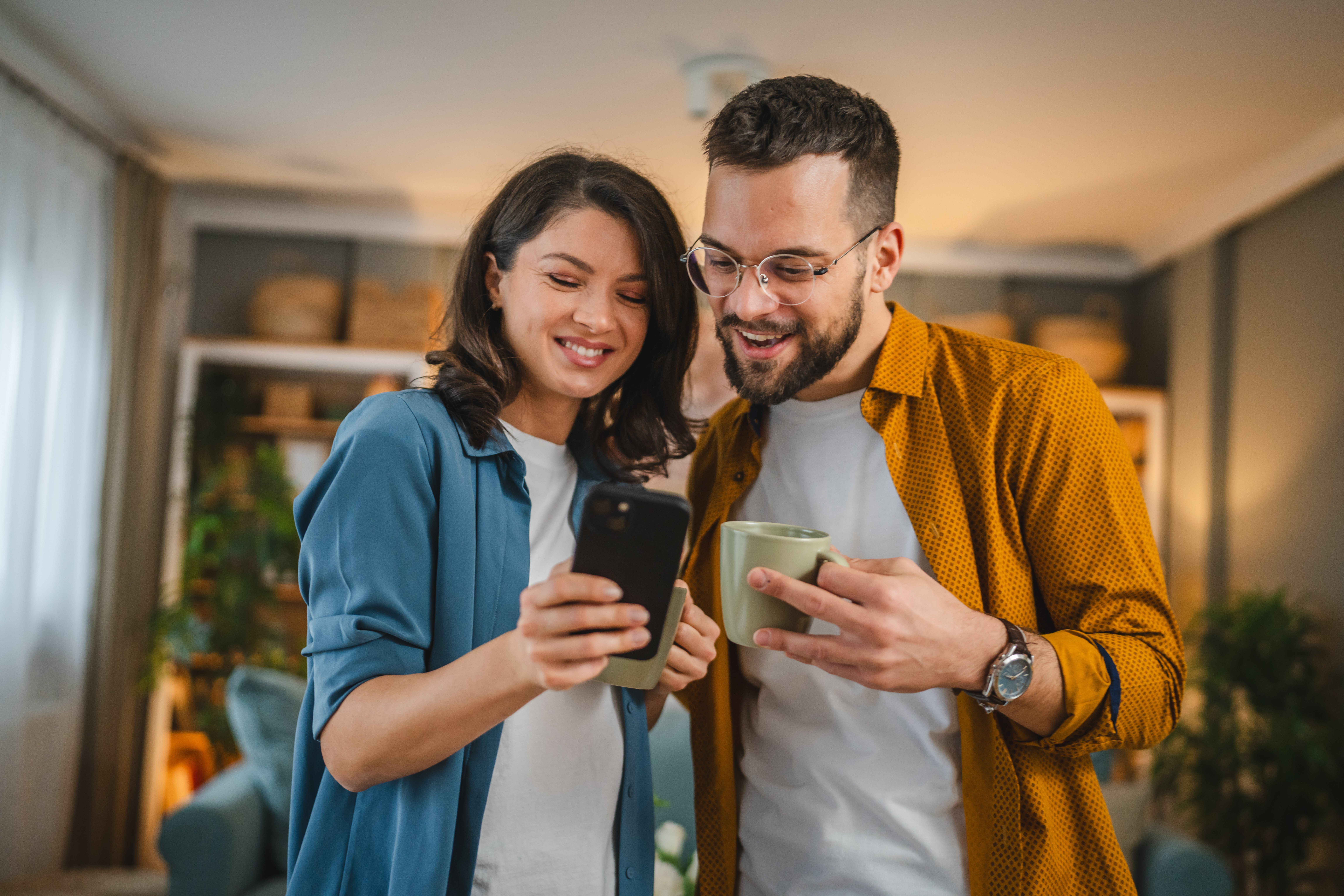 A happy couple enjoying a moment together in a cozy indoor setting, with one person holding a smartphone and the other holding a cup. They both look delighted as they share something on the phone.