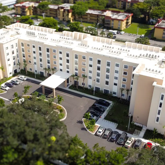 Aerial view of a modern, multi-story hotel building with a light-colored facade surrounded by greenery and a parking lot.