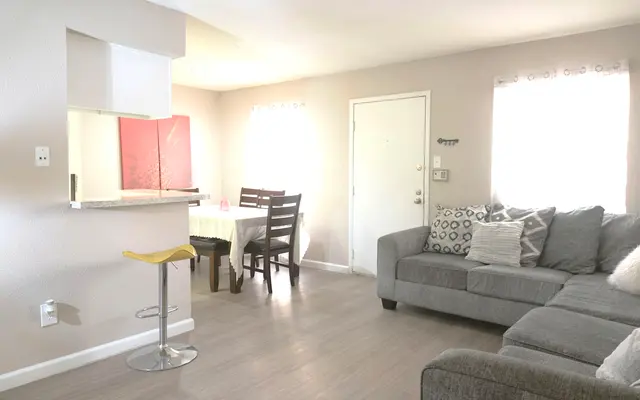 A cozy living room with a grey sectional sofa, decorated with various cushions, a small kitchen area in the background, a dining table with wooden chairs, and a door leading outside.