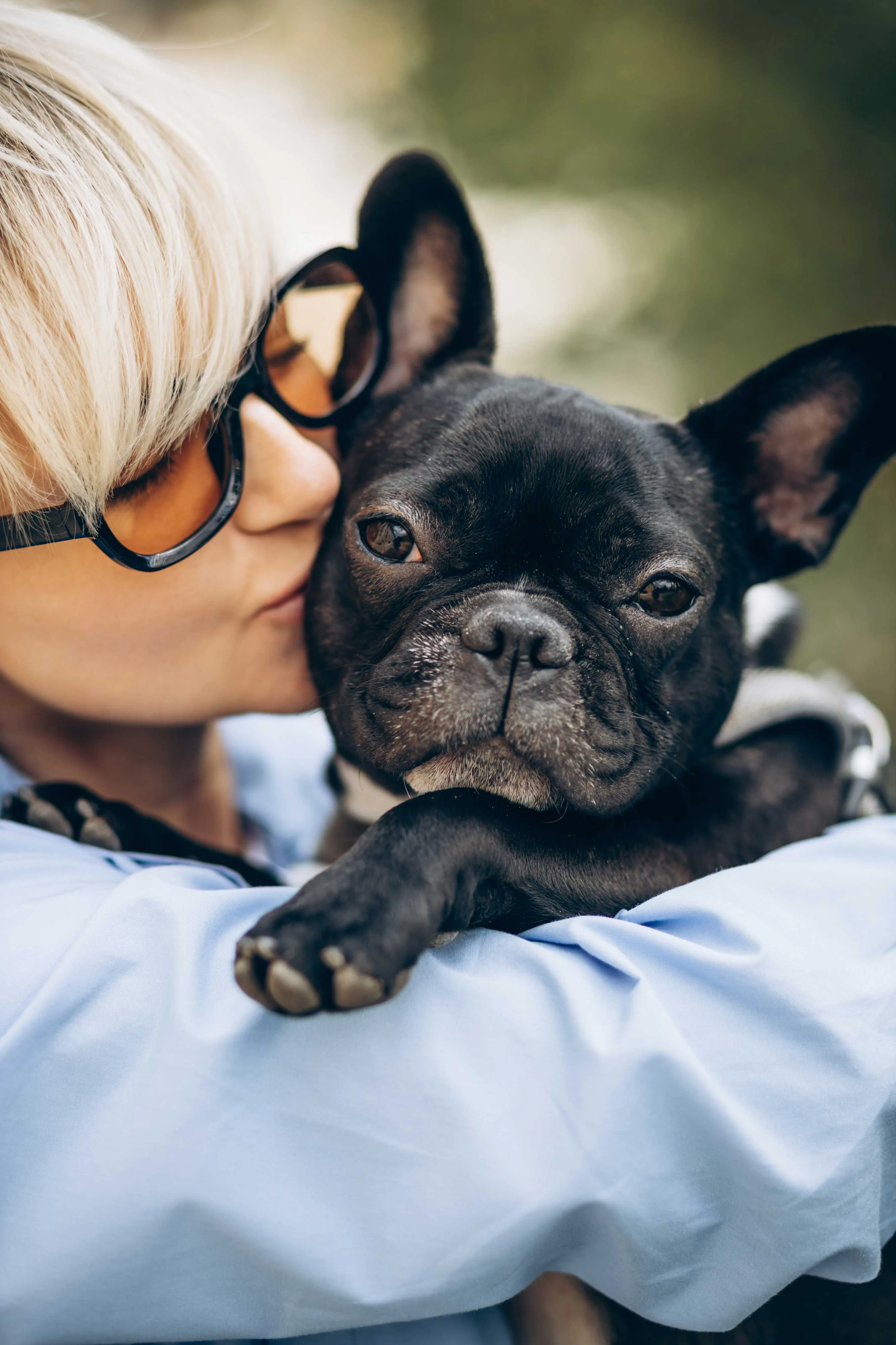 A woman with short blonde hair wearing sunglasses is kissing her French Bulldog, who is being held in her arms. The dog has a calm expression and traits typical of the breed, with a short snout and perked ears. The background is slightly blurred, suggesting a natural outdoor setting.