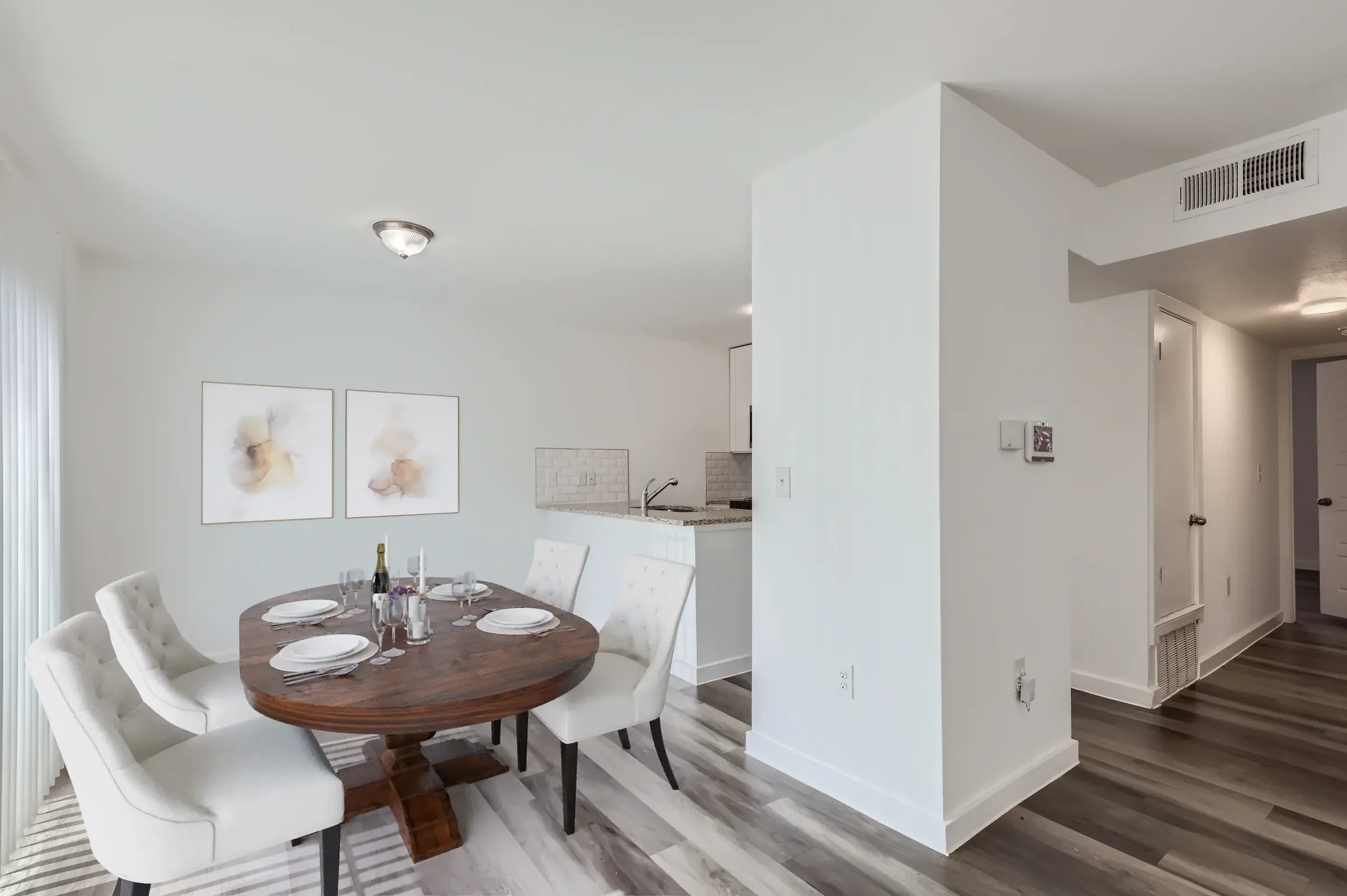 A modern dining area with a round wooden table set for six, featuring white upholstered chairs. There are two abstract framed artworks on the wall and a glimpse of a kitchen in the background. The floor has a mixed wood design.