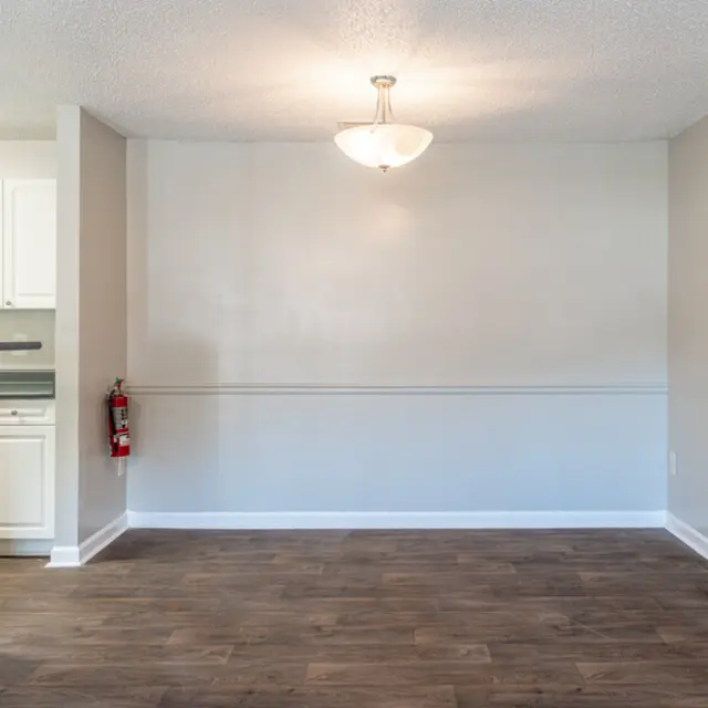 An empty living room space with wooden flooring, a wall-mounted light fixture, and a view of a kitchen area with white cabinetry in the background.