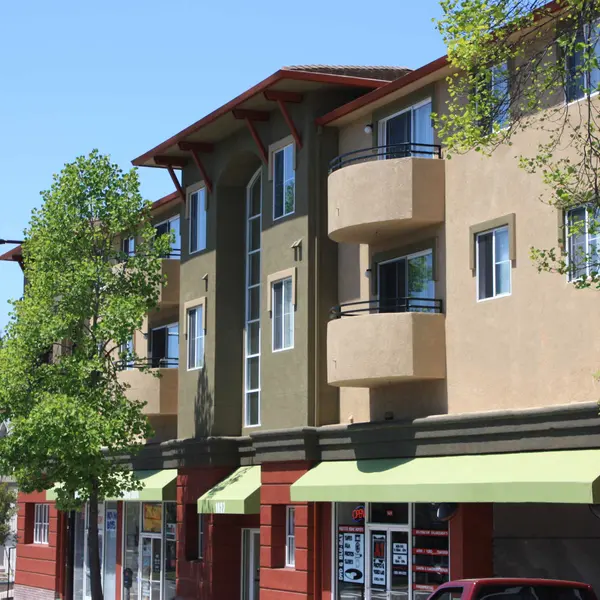 A modern apartment building with a mix of beige and green facades, featuring balconies and large windows. The ground level has shops with green awnings.
