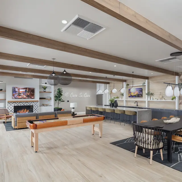 A spacious modern common area with wooden beams, large dining tables, and a cozy seating area featuring a fireplace and decorative plants.