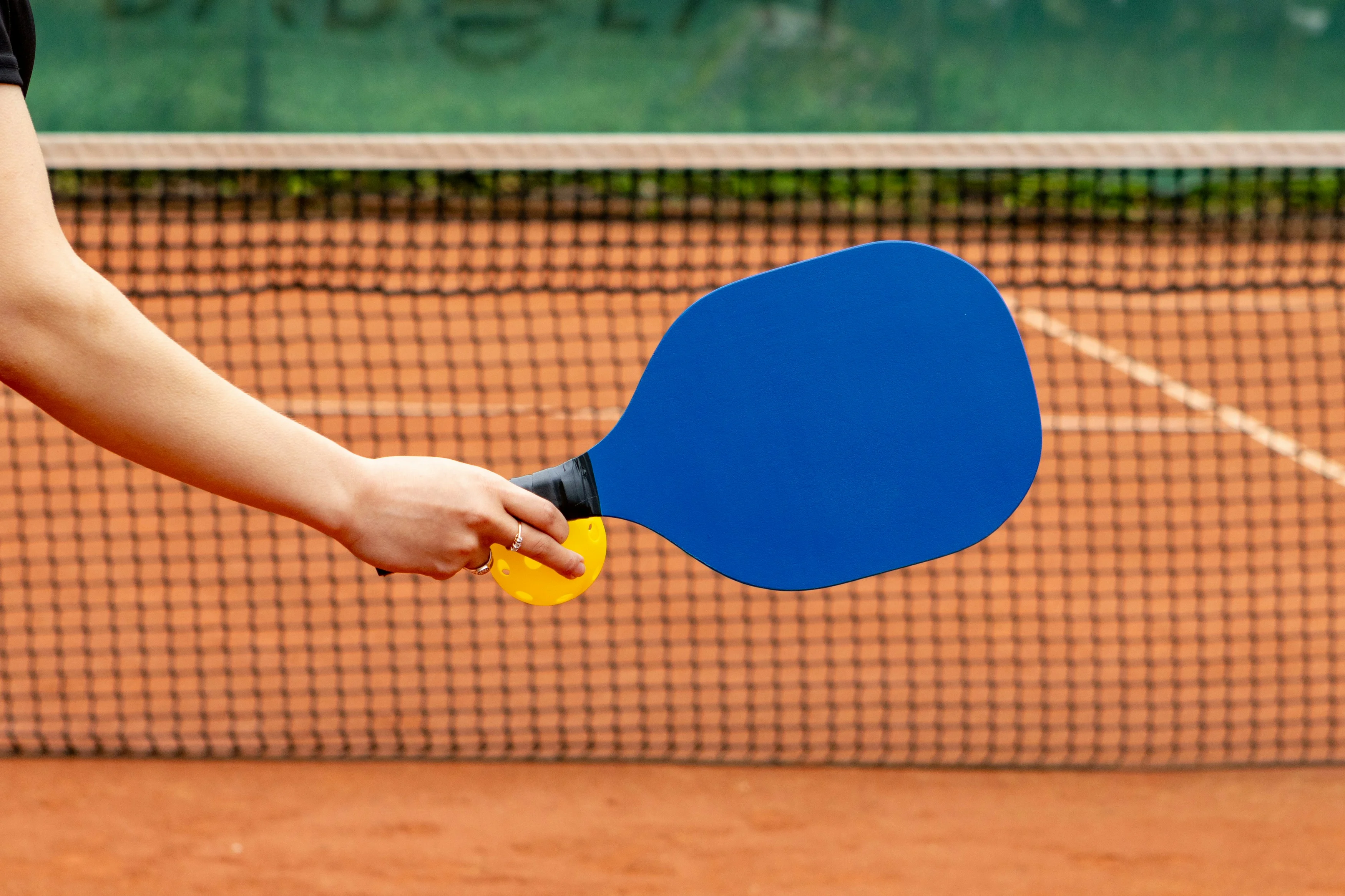 Paddle Sport Action Shot A person holding a blue paddle with a yellow grip, standing on a clay court near a net.