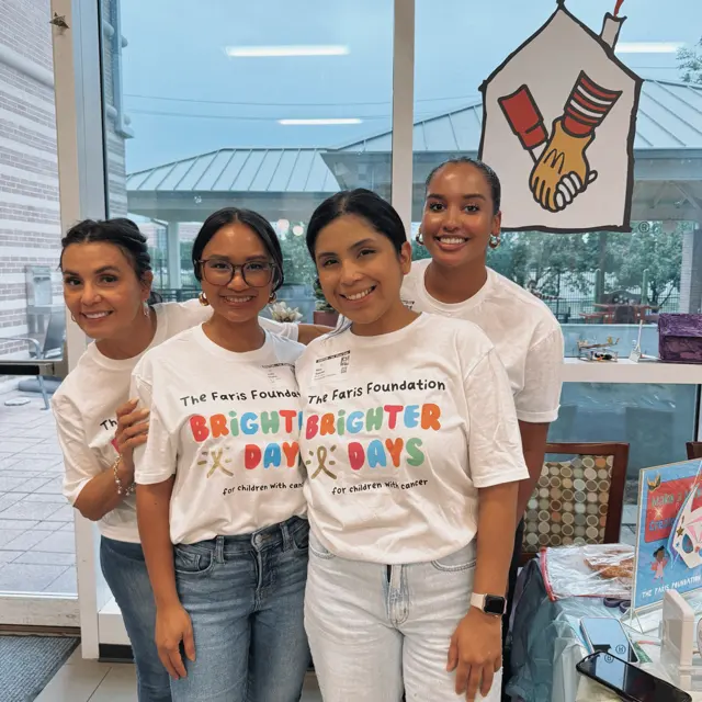 Brighter Days Community Event A group of four individuals wearing matching t-shirts that say 'Brighter Days' and 'The Faris Foundation.' They are smiling and posing together in a community setting with a drawing of a heart and hands in the background.