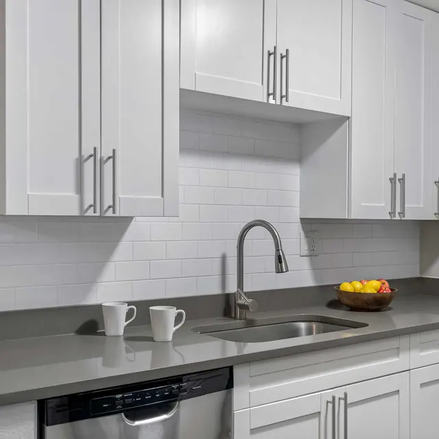 A modern kitchen with white cabinetry and a gray countertop, featuring a sink, a dishwasher, and a bowl of fruit on the counter.