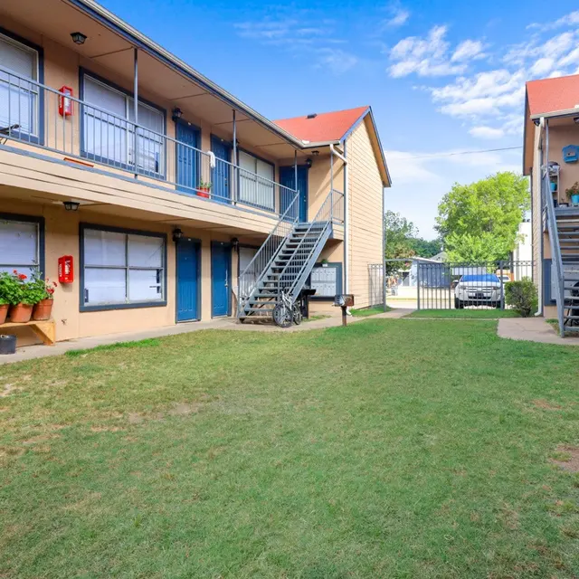A courtyard view of a two-story apartment complex featuring a green lawn, flower pots, and staircases leading to the upper units.