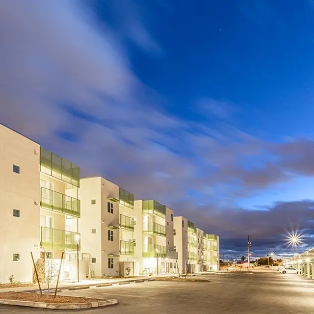 A modern apartment complex with a spacious parking area under a cloudy evening sky. The buildings are white with green accents, and the scene is illuminated by streetlights.