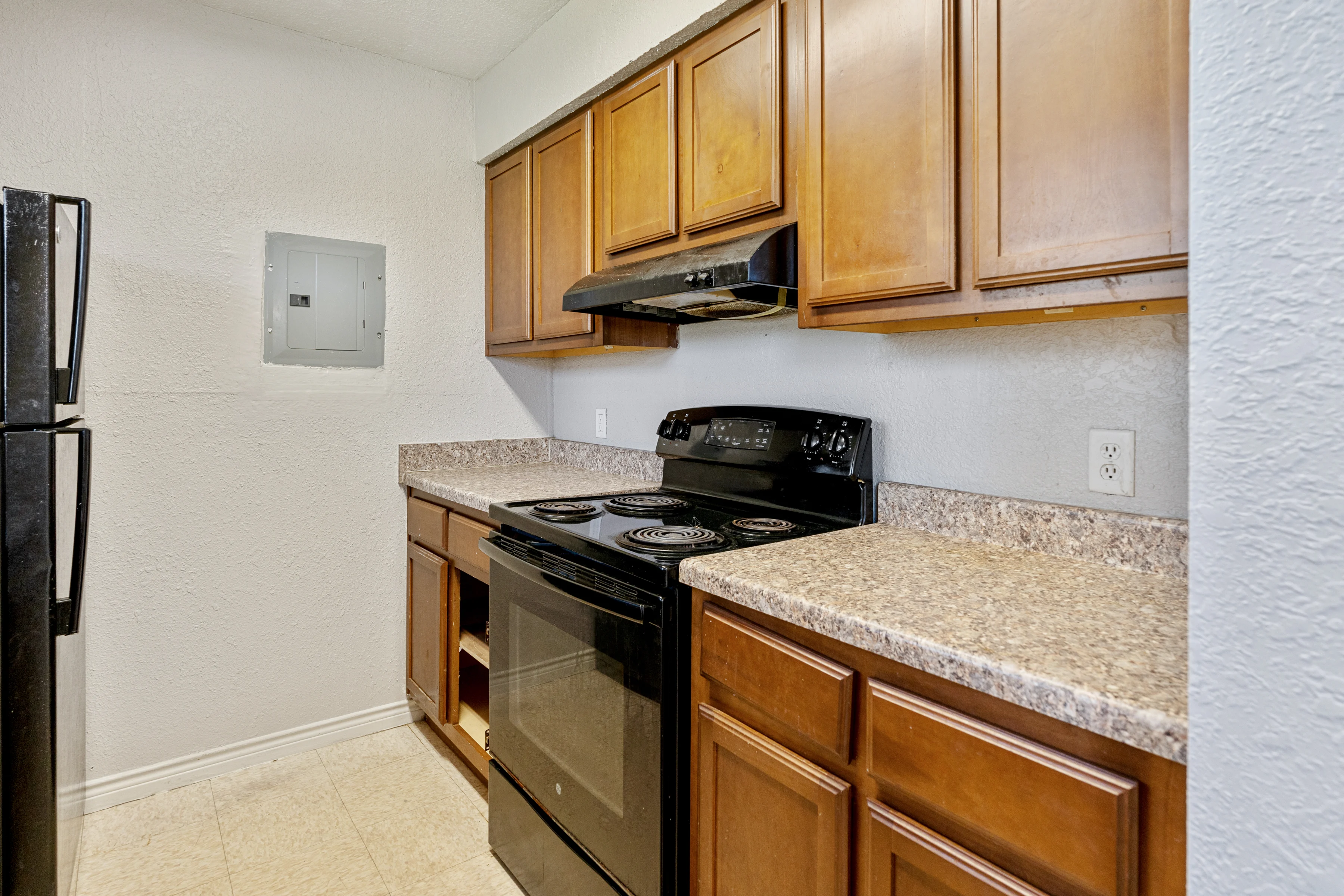 Compact Kitchen Interior A small kitchen featuring wooden cabinets, a black stove and oven, a granite-like countertop, and a black refrigerator. An electrical panel is visible on the wall.