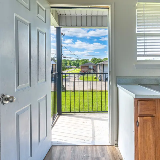 Inside an apartment showing an open door leading to a balcony with a view of green grass and a blue sky.