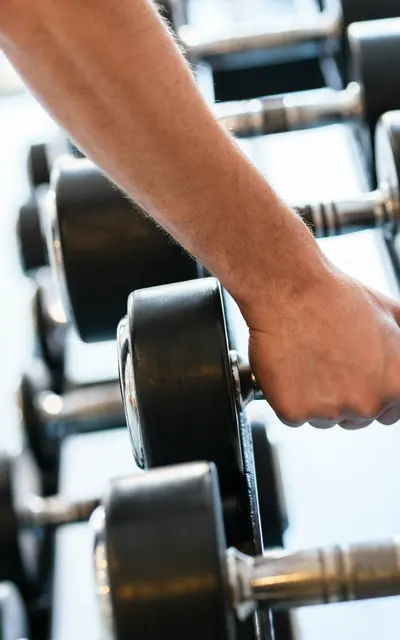 Bayshore Pines A close-up of a hand gripping a dumbbell in a gym setting, with multiple dumbbells lined up in the background.