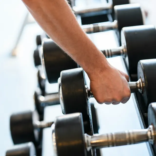 A close-up of a hand gripping a dumbbell in a gym setting, with multiple dumbbells lined up in the background.
