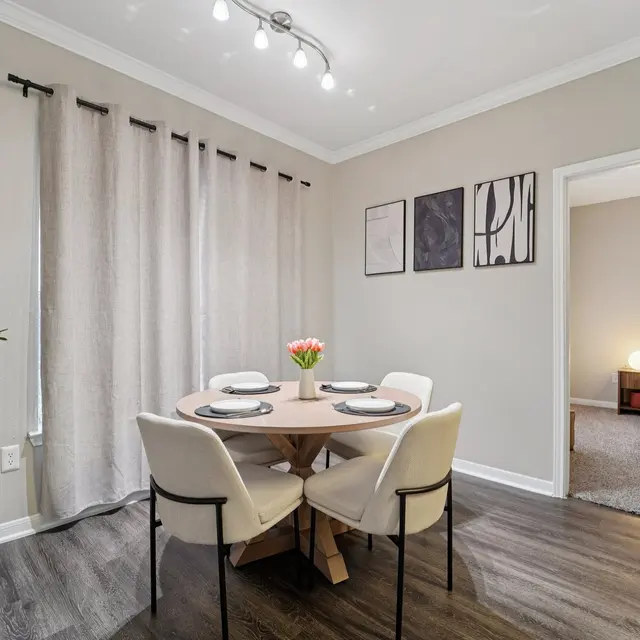 A modern dining area featuring a round wooden table with four cream-colored chairs, a small vase of flowers on the table, and a large window with light gray curtains. A decorative plant is in the corner. In the background, a doorway leads to a bedroom with soft lighting.