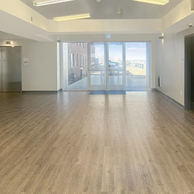 A spacious modern lobby area with wood flooring and large glass doors opening outside. There are elevators on the left wall and a solid black door on the right.