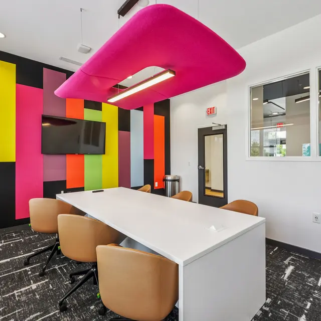 Modern Meeting Room with Colorful Design A colorful modern meeting room featuring a long white table and brown chairs. The walls are adorned with vertical panels in vibrant colors, and a large pink acoustic panel hangs from the ceiling. A television is mounted on one wall.