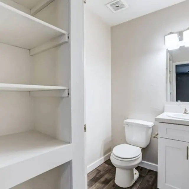 A modern bathroom with a white vanity, toilet, and open shelving space on one side.