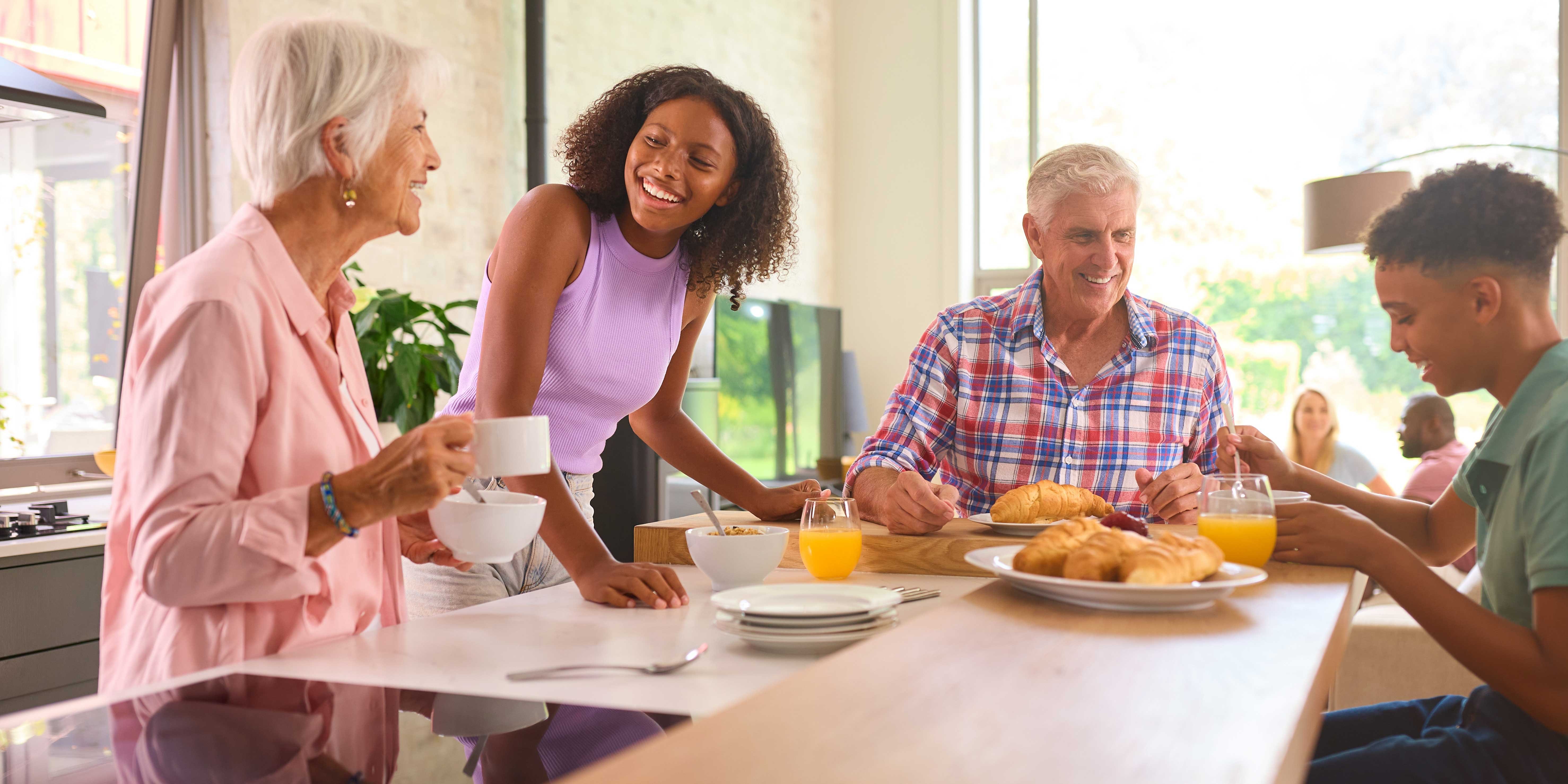 Family Breakfast Gathering A family enjoying breakfast in a bright, modern kitchen, featuring an elderly woman, a young woman, a middle-aged man, and a teenage boy, all smiling and interacting over food and drinks.