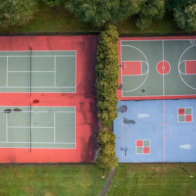Aerial view of two sports courts side by side; the left side features two tennis courts with a green surface, and the right side showcases a basketball court with a blue surface.