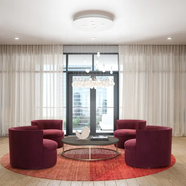 A modern living room featuring a circular arrangement of four plush purple chairs around a glass coffee table on a round red area rug. The background consists of large windows with sheer drapes and dark wooden shelves showcasing decorative items.