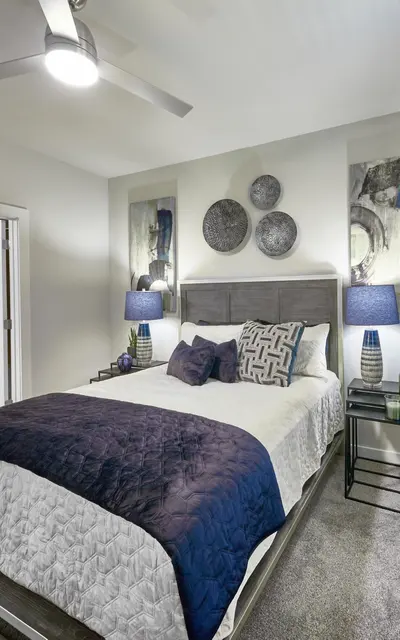 A modern bedroom featuring a queen bed with a navy blue and white color scheme, two bedside tables with lamps, and artwork on the walls. A door leads to another room while a window lets in natural light.