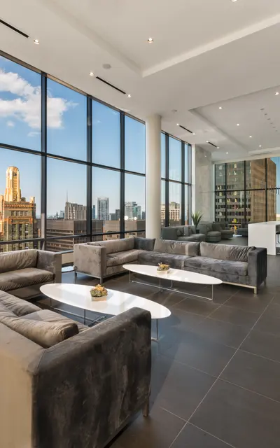 A modern living room with large windows offering a view of a city skyline. The room features contemporary furniture, including gray sofas, a white coffee table, and a sleek design with a focus on natural light.