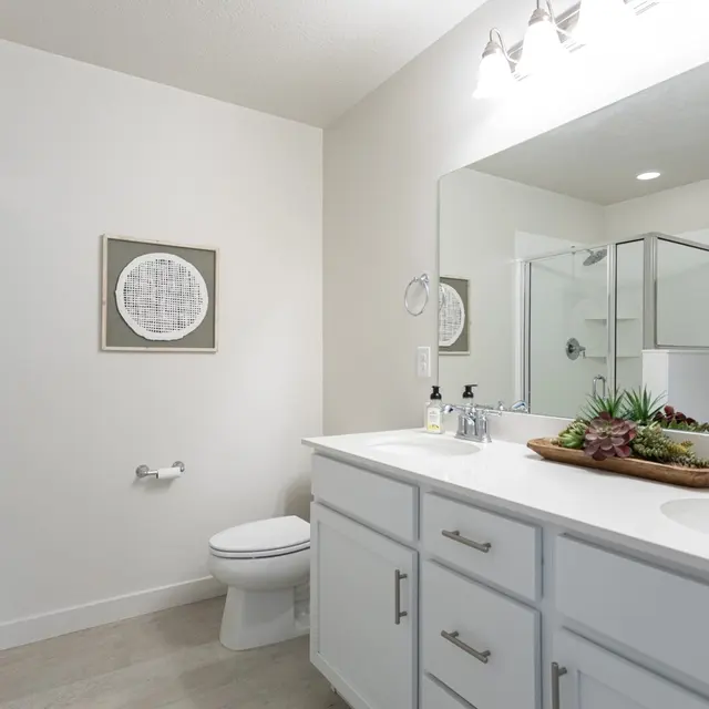 A modern bathroom featuring a white double vanity with a large mirror, a minimalist decor piece on the wall, a shower area behind glass, and neutral-colored flooring.