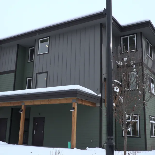 A modern apartment building with a combination of green siding and wooden features, surrounded by snow on the ground and some leafless trees in winter.