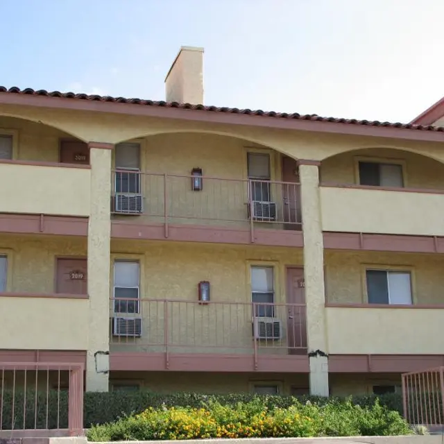 Apartment Building Exterior Exterior view of a multi-story apartment building with three levels, featuring yellow walls and light pink accents. Balconies with railings are present on each level, and several windows are visible. The building is surrounded by greenery and flowers.
