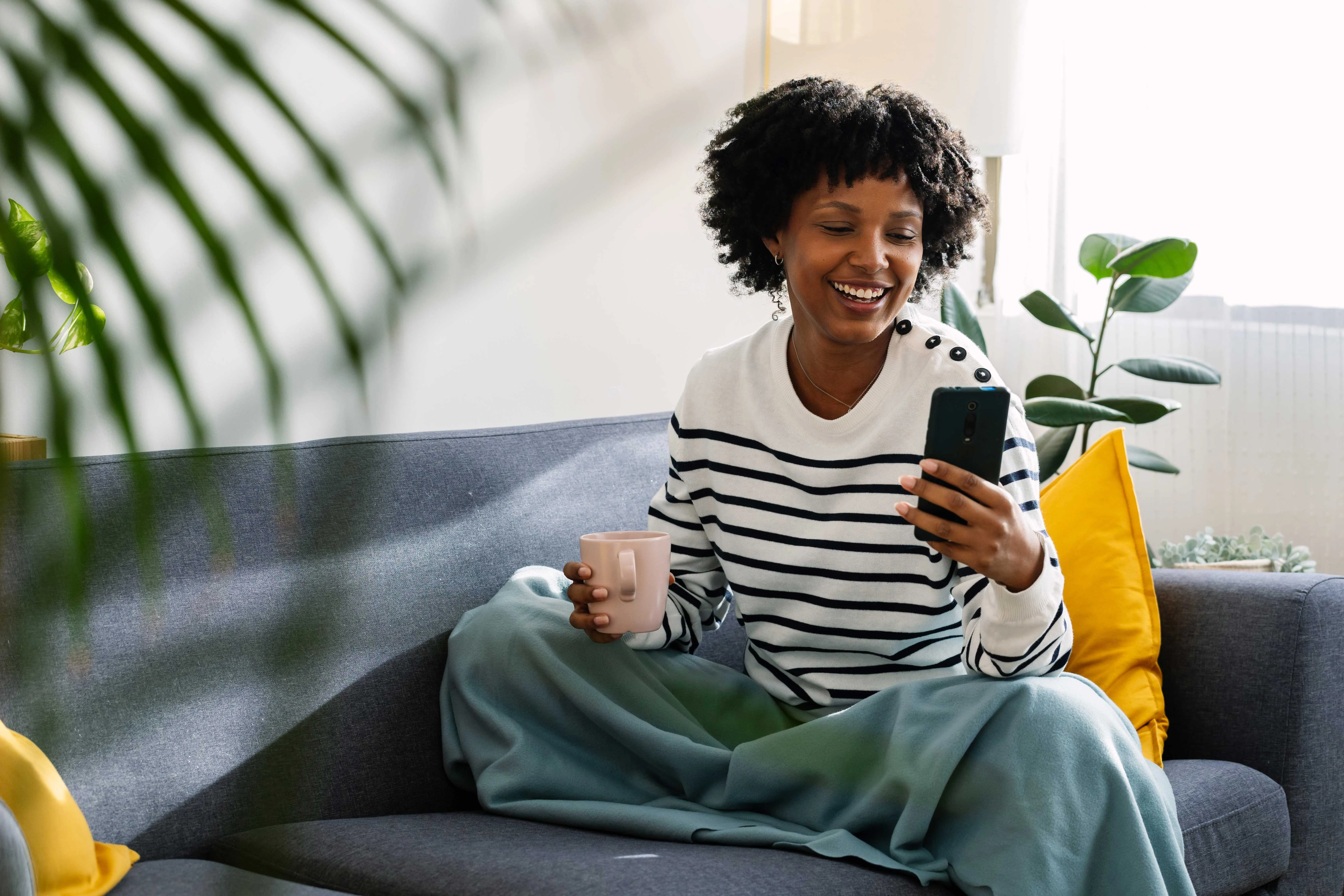 A woman sitting on a couch with her legs crossed, holding a phone in one hand and a mug in the other, smiling at her phone. Bright and airy living room with plants and decorative pillows in the background.