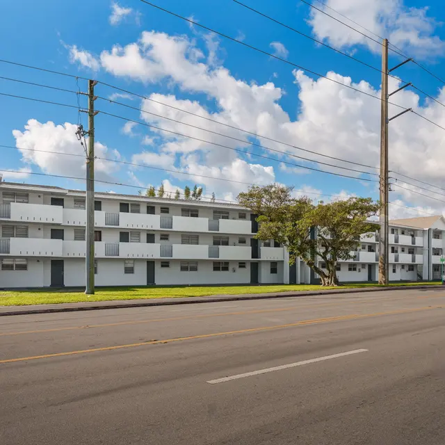 A view of a modern apartment building from across the street, showing multiple floors and balconies under a partly cloudy blue sky.