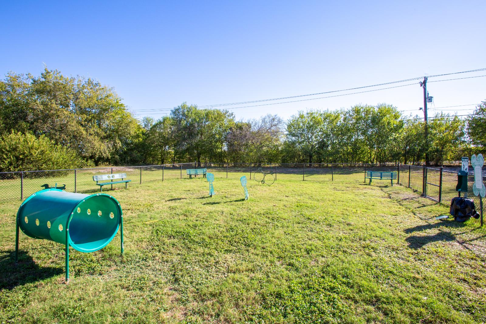 An open area of a dog park featuring green play equipment and benches surrounded by trees and a fence.