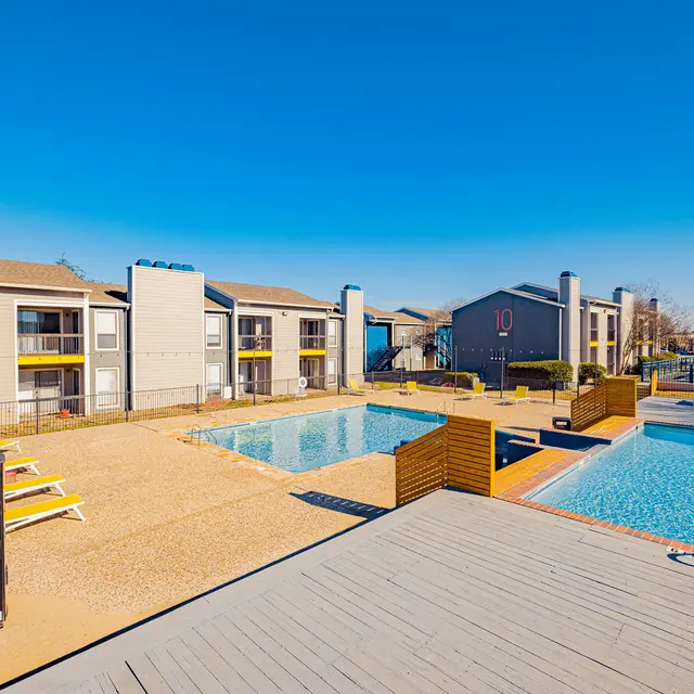 A view of a swimming pool area surrounded by apartment buildings, featuring lounge chairs and a pool deck.