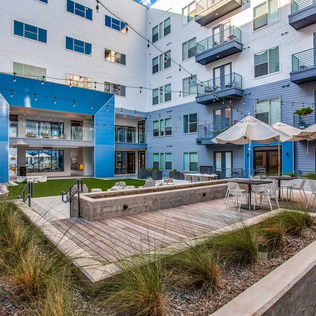 A modern courtyard in an apartment complex with grass, lounge chairs, and barbecue facilities. There are umbrellas over seating areas and multiple balconies visible from surrounding units.