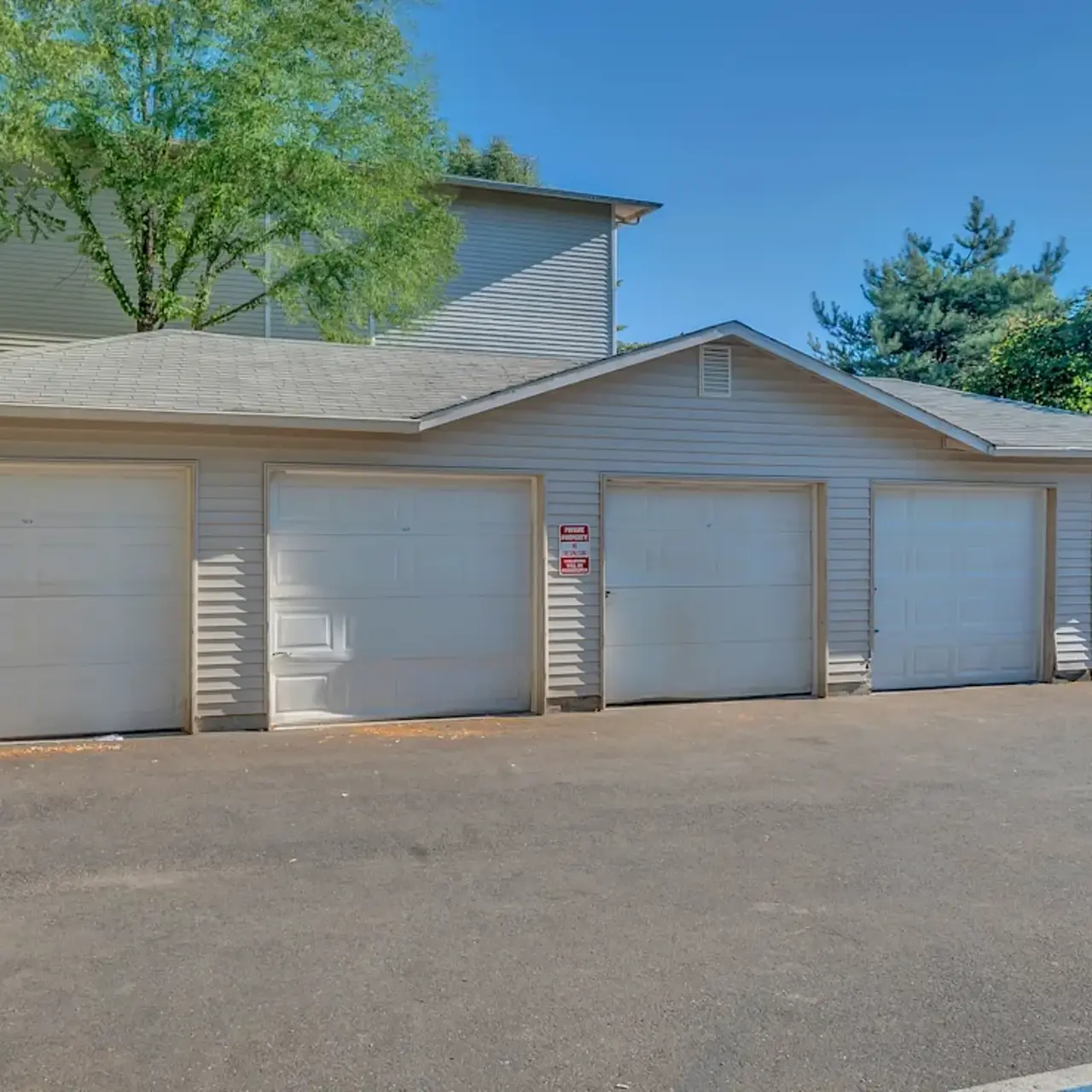 A large multi-door garage with four closed white doors, surrounded by trees and shrubs under a clear blue sky.