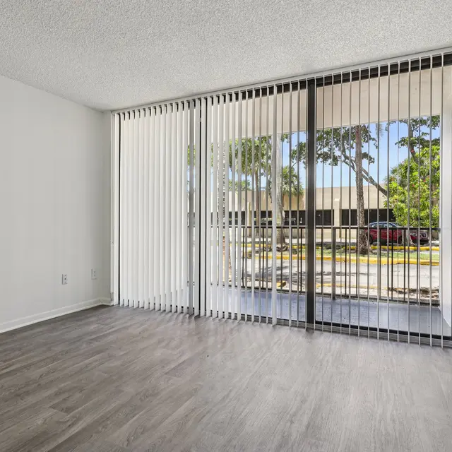 Bedroom - Biscayne Apartments An empty room featuring sliding glass doors with vertical blinds, overlooking an outdoor area with trees and a parking lot. The flooring is light wood laminate and the walls are painted white.