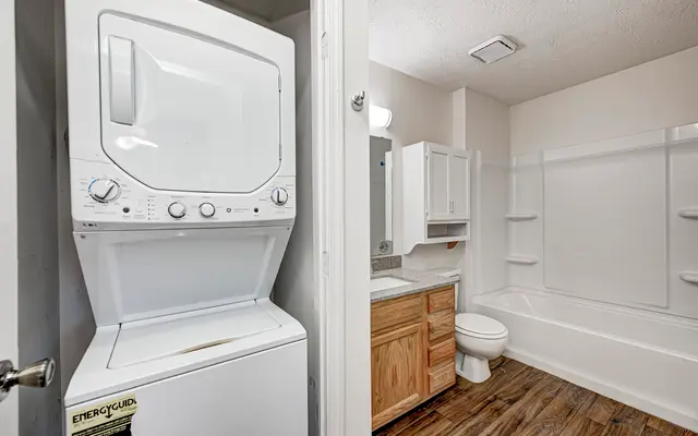 A laundry appliance located in a bathroom, featuring a stacked washer and dryer on the left side and a bathtub with a shower, toilet, and wooden vanity on the right side.