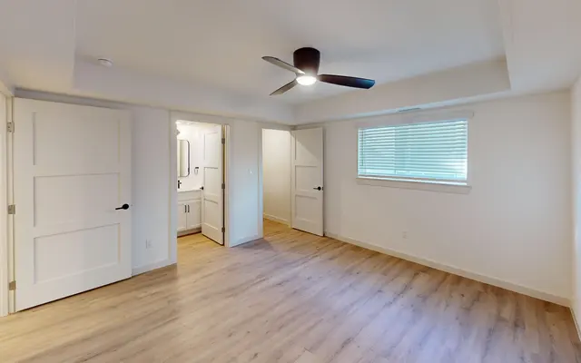 A spacious, modern bedroom with light wood flooring, featuring a ceiling fan and two closed doors leading to other areas. Natural light enters through a window with blinds.