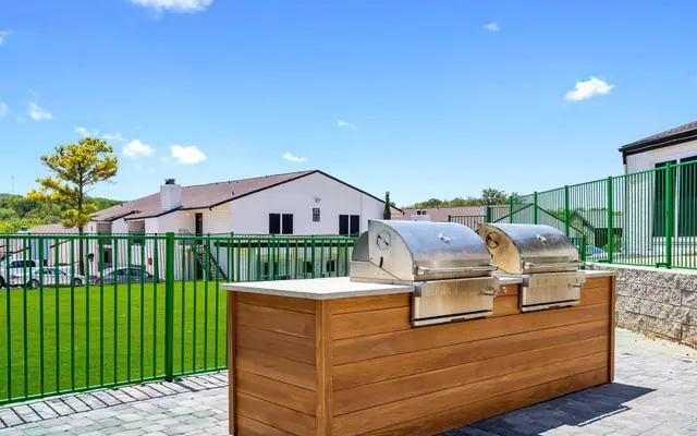 An outdoor grilling area featuring two stainless steel grills on a wooden counter, with a green lawn and fence in the background under a clear blue sky.