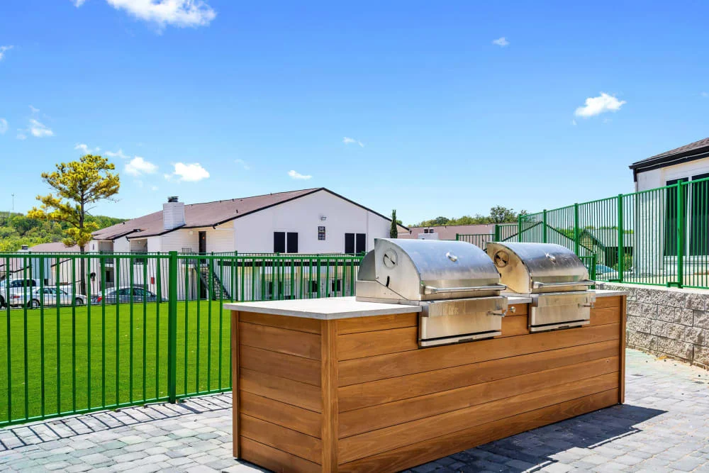 An outdoor grilling area featuring two stainless steel grills on a wooden counter, with a green lawn and fence in the background under a clear blue sky.
