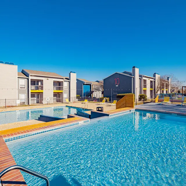 A clear blue swimming pool in an apartment complex, with sun loungers and buildings in the background under a bright blue sky.